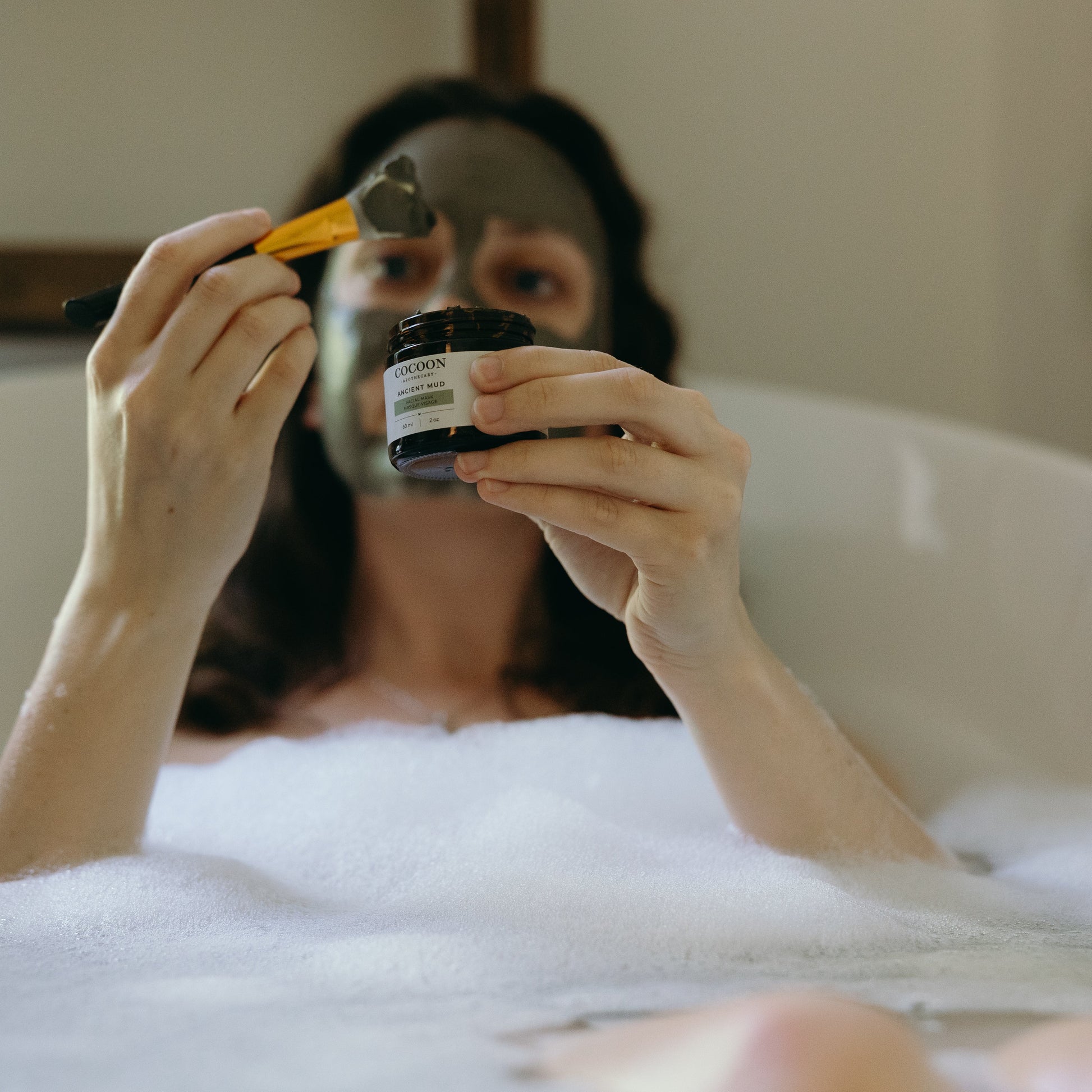 Person in a bubble bath holding Ancient Mud and a brush.