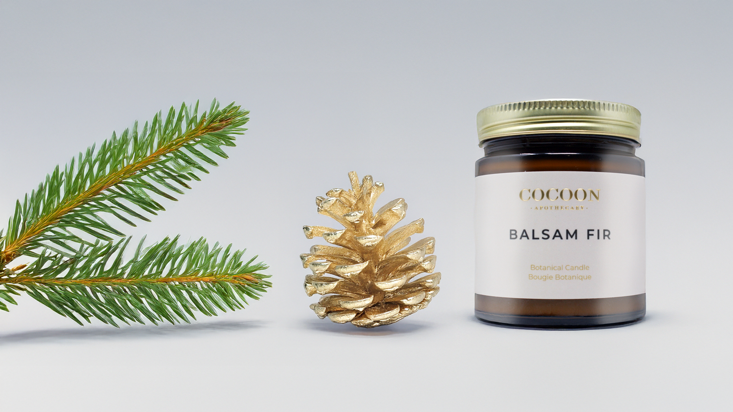 Cocoon Balsam Fir candle with a pine branch and gold pine cone on a light gray background