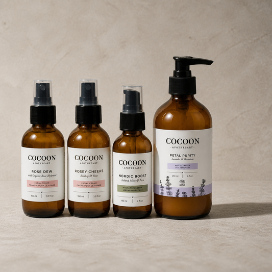 Four Cocoon Apothecary skincare bottles in amber glass with black pumps and white labels, arranged on a light beige stone surface against a textured beige wall. The bottles include Rose Dew, Rosey Cheeks, Nordic Boost, and Petal Purity.