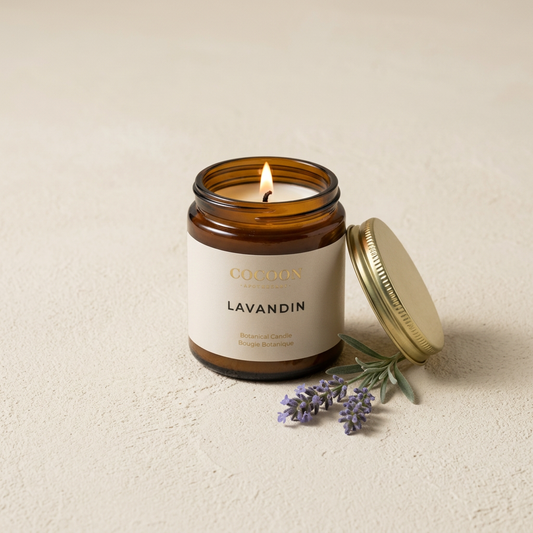 An amber glass jar candle labeled 'COCOON LAVANDIN' with a lit flame, shown with its gold lid beside it and small lavender sprigs on a light beige textured surface.