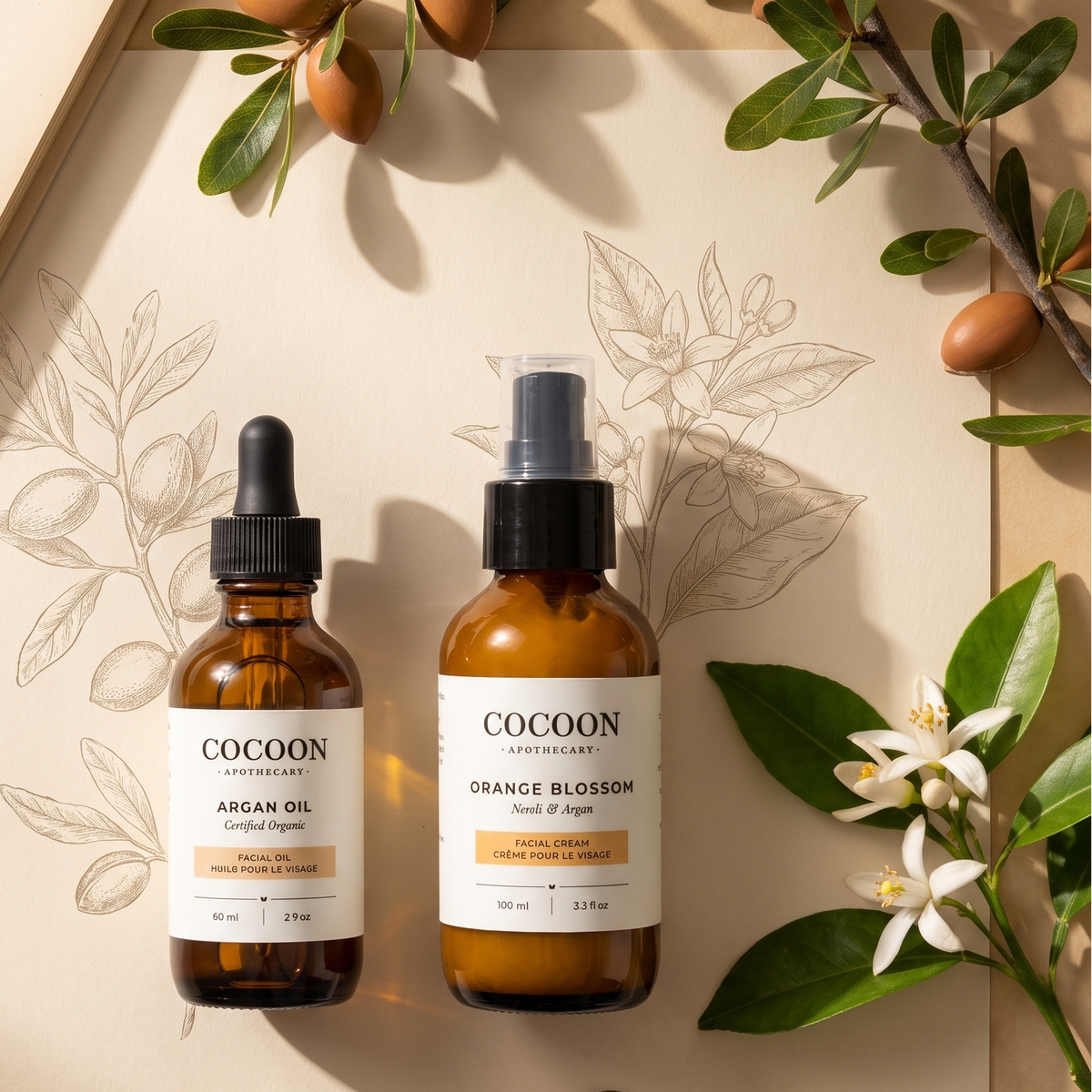 Two amber glass skincare bottles from Cocoon Apothecary one with a dropper and one with a spray, arranged on a beige paper background with botanical illustrations, an open book, a vintage key, and citrus leaves and flowers.