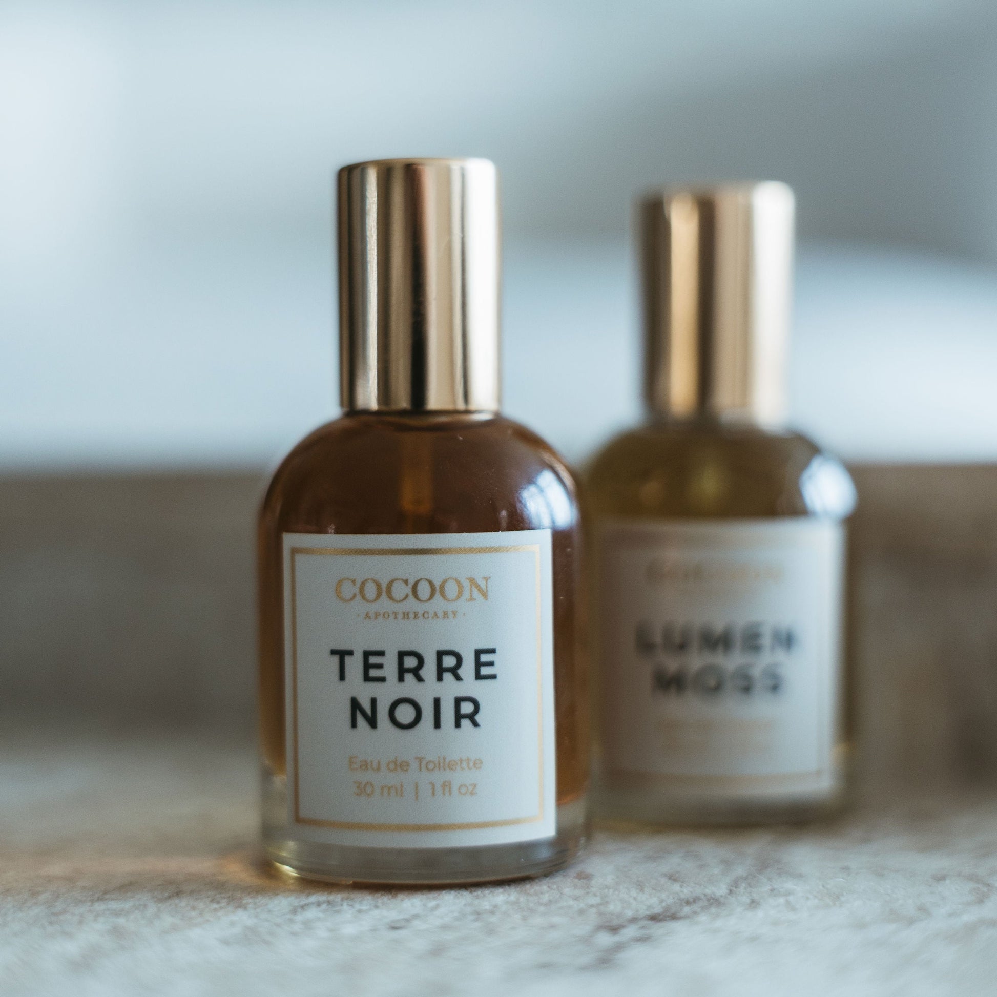 Two perfume bottles labeled 'Terre Noir' and 'Lumen Moss' on a marble surface with a blurred background.