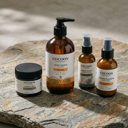 Four Cocoon Apothecary skincare and products arranged on a gray stone surface: a large pump bottle of Sweet Orange body wash, a smaller bottle of Nordic Boost serum, a bottle of Orange Blossom cream, and a jar of Ancient Mud. The bottles are amber glass with black caps and white labels.